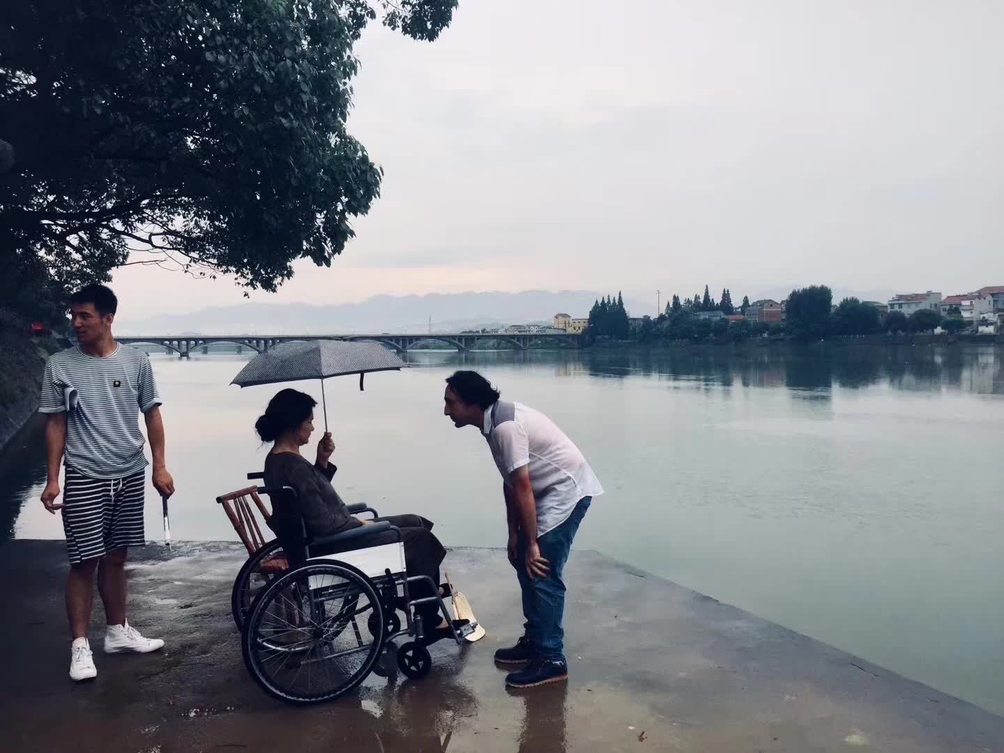 三人在河边，其中一人坐在轮椅上，撑着雨伞。
